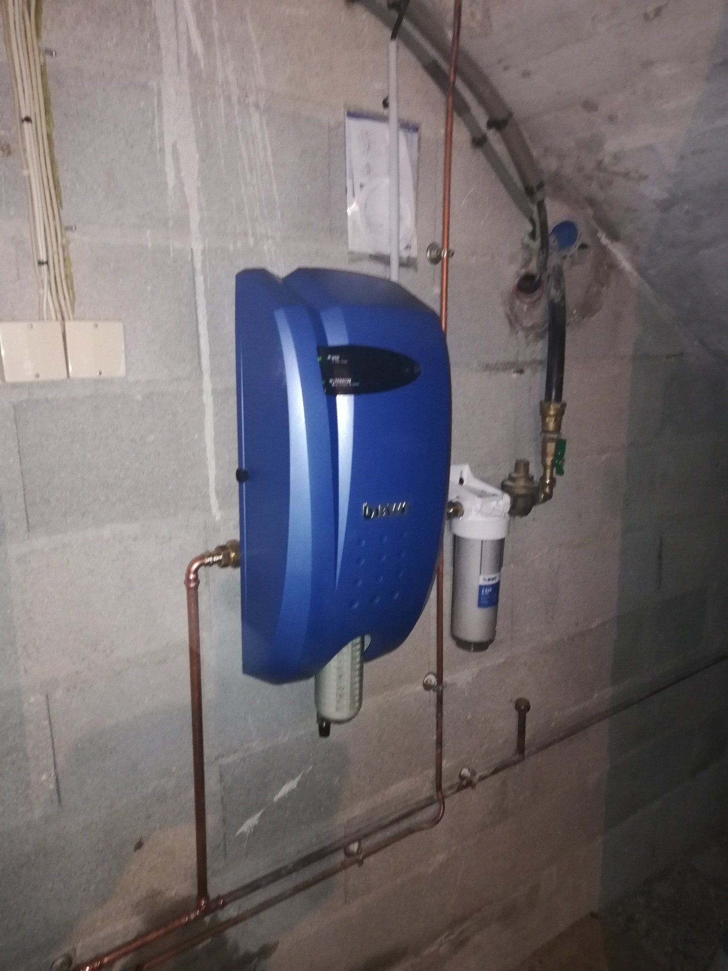 Un adoucisseur d'eau bleu fixé sur un mur en parpaings, relié à des tuyaux en cuivre et à un filtre.