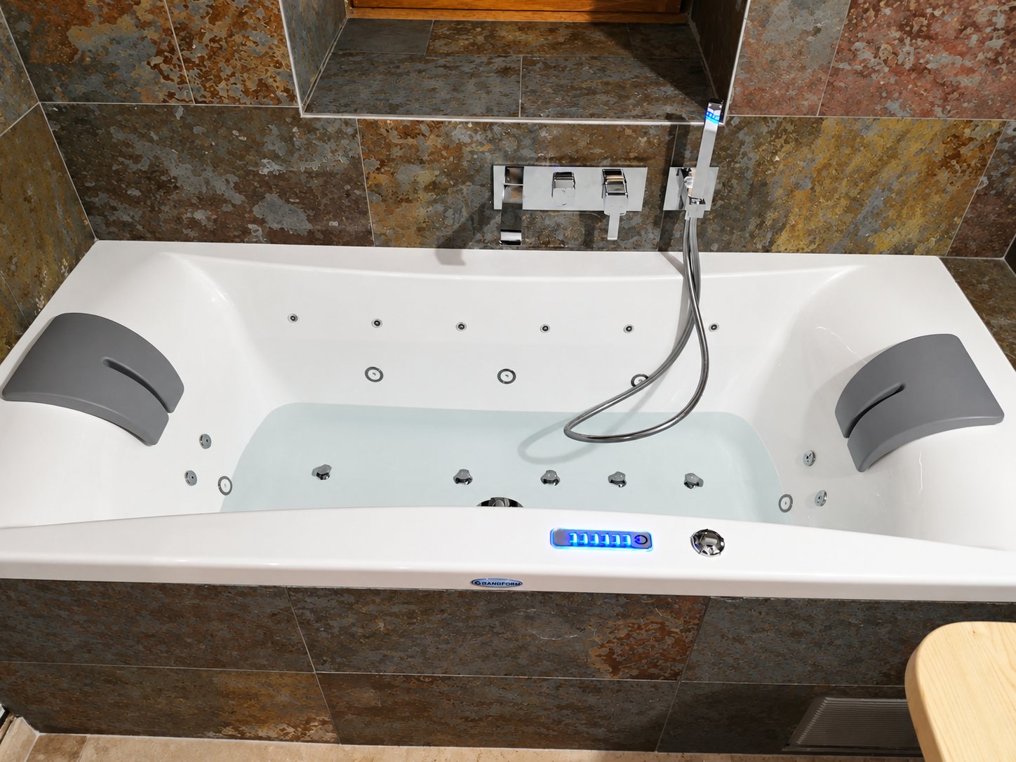 Baignoire balnéo blanche à deux places avec jets, appuie-tête gris, douchette et commande lumineuse bleue, encastrée dans un habillage de carrelage effet ardoise.