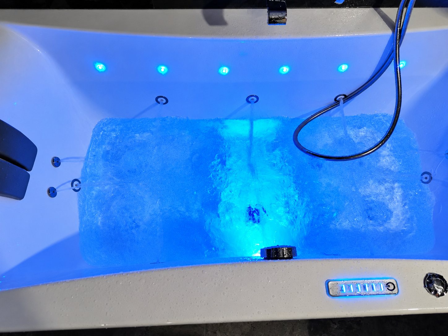 Vue du dessus d'une baignoire balnéo en marche, éclairée par des spots bleus, avec jets d'eau, douchette et panneau de commande tactile.