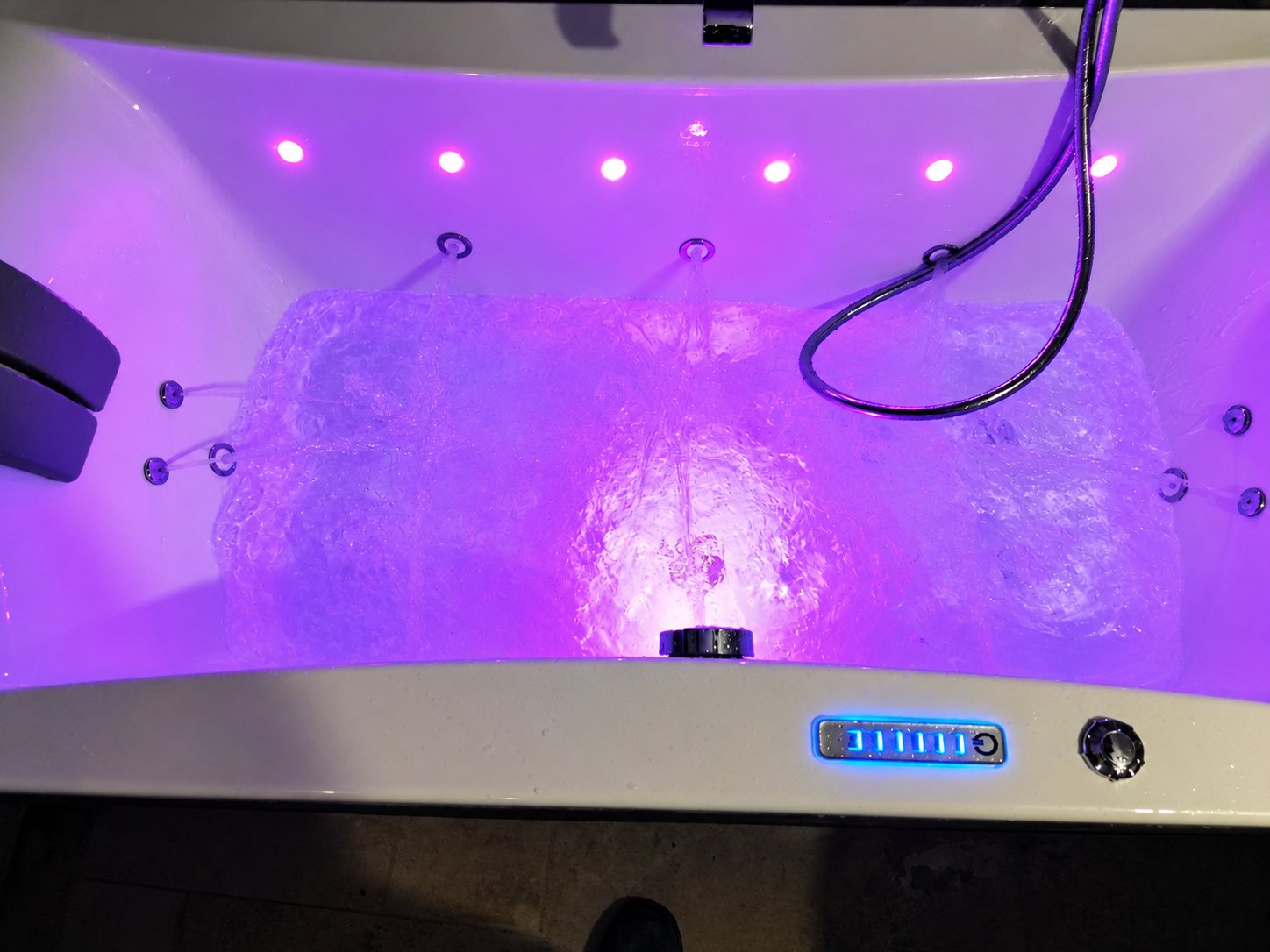 Vue de dessus d'une baignoire balnéo remplie d'eau, éclairée en violet par des spots LED, avec ses jets, sa douchette et son panneau de commande tactile bleu.