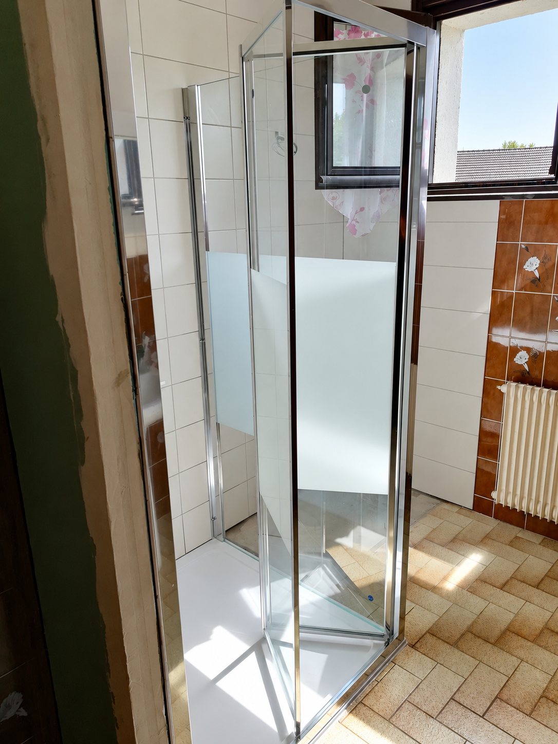 Une cabine de douche à profilés chromés et verre transparent avec bande dépolie, installée dans une salle de bain carrelée avec un radiateur et une fenêtre.