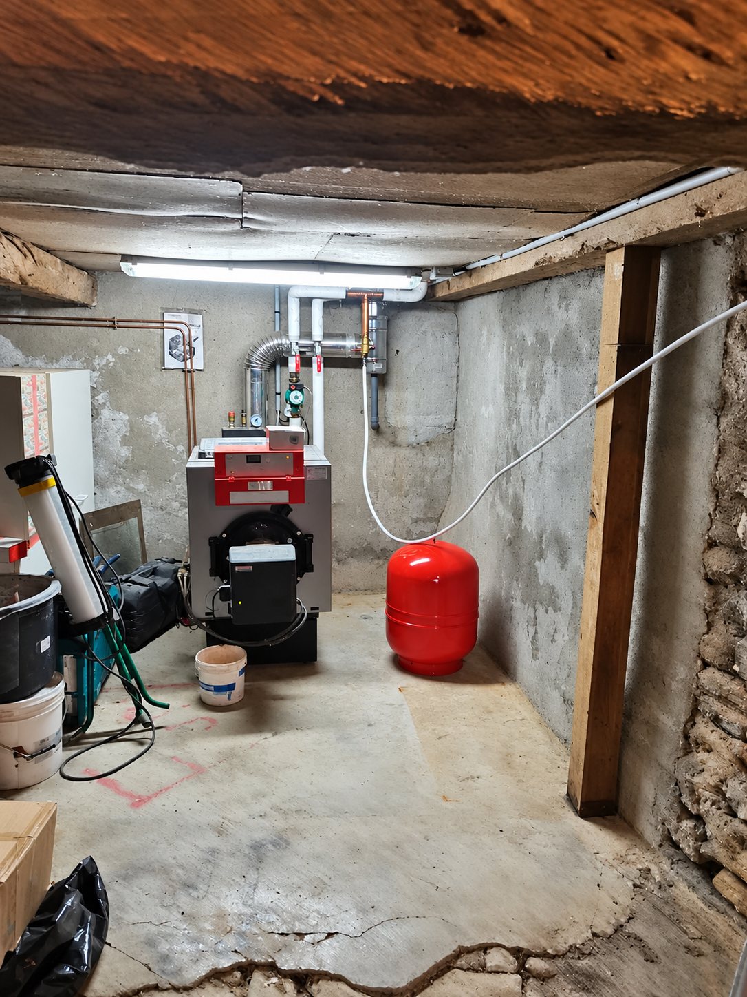 Une chaudière avec brûleur rouge et un vase d'expansion rouge, raccordés par des tuyaux, installés dans une cave en pierre éclairée par un néon.