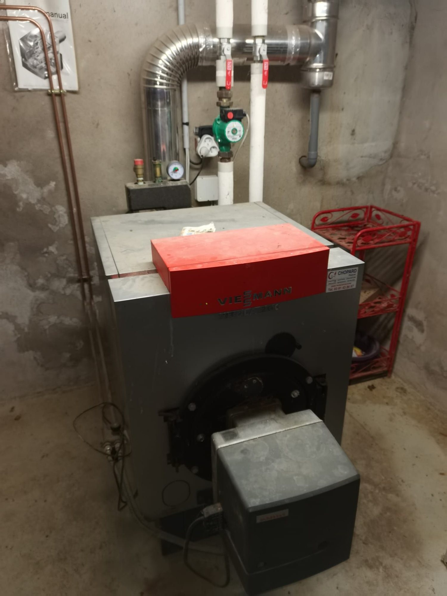 Une chaudière Viessmann grise et rouge équipée d'un brûleur est installée dans un local technique en béton, avec tuyaux de cuivre, conduit de fumée en inox et circulateur vert.