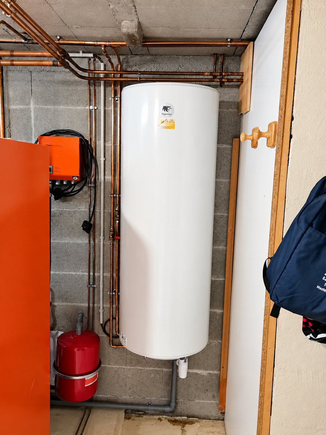 Un ballon d'eau chaude blanc de marque Thermor est fixé au mur de parpaings d'un local technique, entouré de tuyaux en cuivre et d'un vase d'expansion rouge.