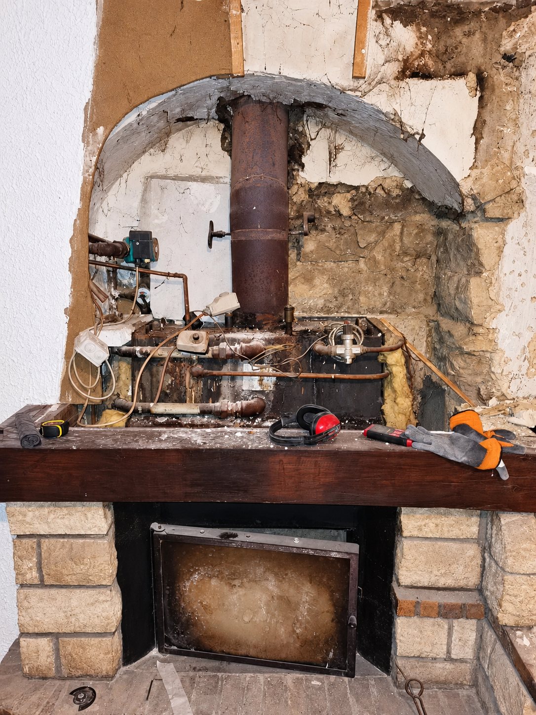 Une cheminée en pierre en cours de rénovation, avec une chaudière ancienne et des tuyauteries visibles derrière le manteau en bois où sont posés un casque antibruit et des gants.