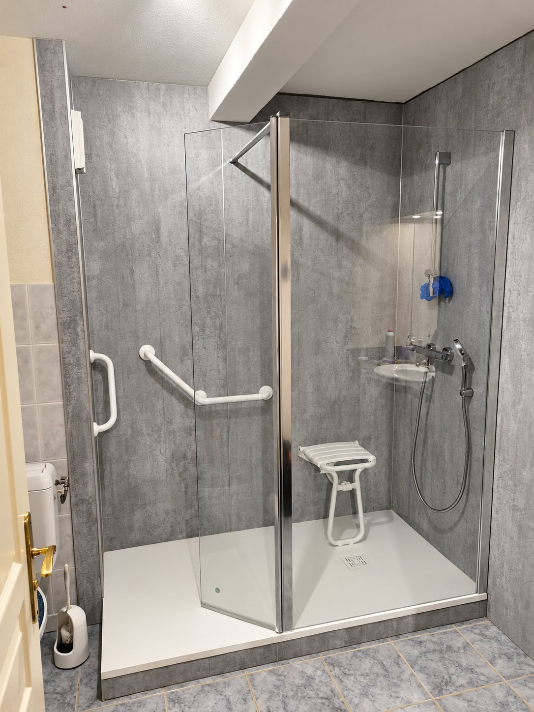 Une douche rénovée aux parois grises effet béton, avec receveur blanc, porte vitrée, siège rabattable et barres d'appui blanches.