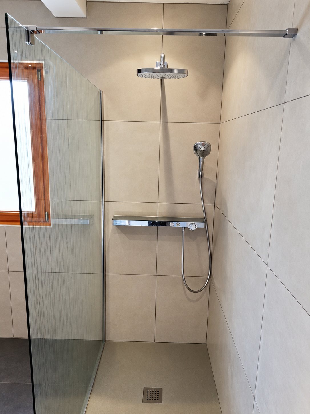 Douche à l'italienne aux grands carreaux beiges, avec pomme de pluie, douchette, étagère thermostatique et paroi de verre.