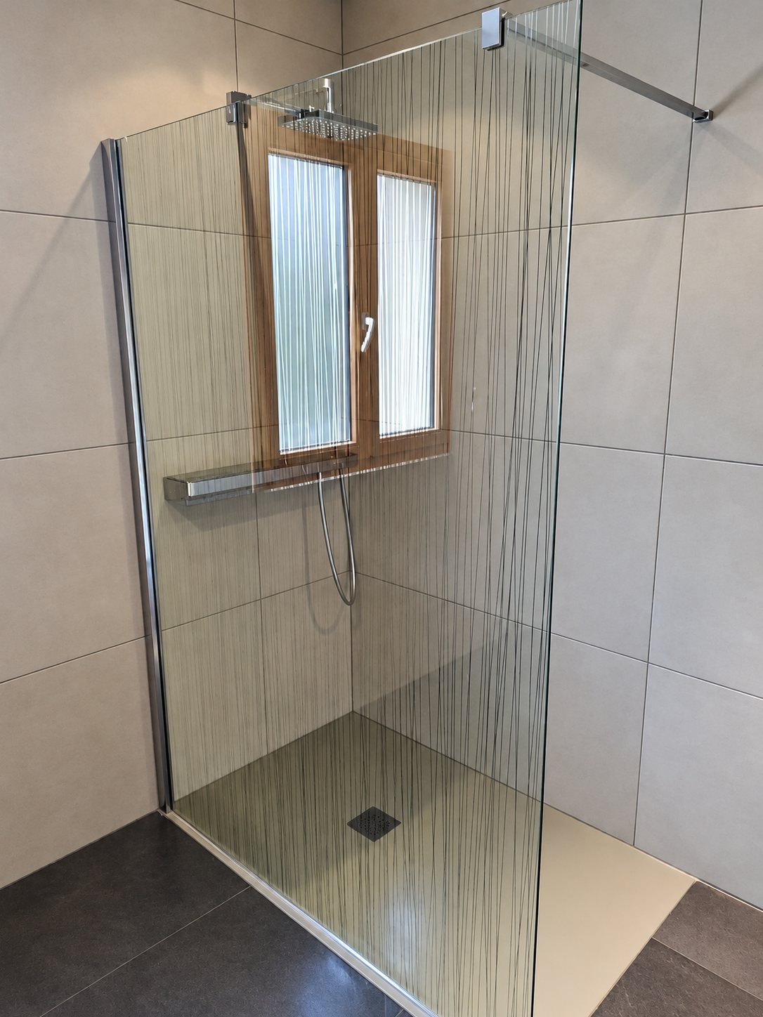 Une douche à l'italienne carrelée de grands carreaux gris, fermée par une paroi de verre à fins traits noirs, avec une fenêtre et une pomme de douche de style pluie.