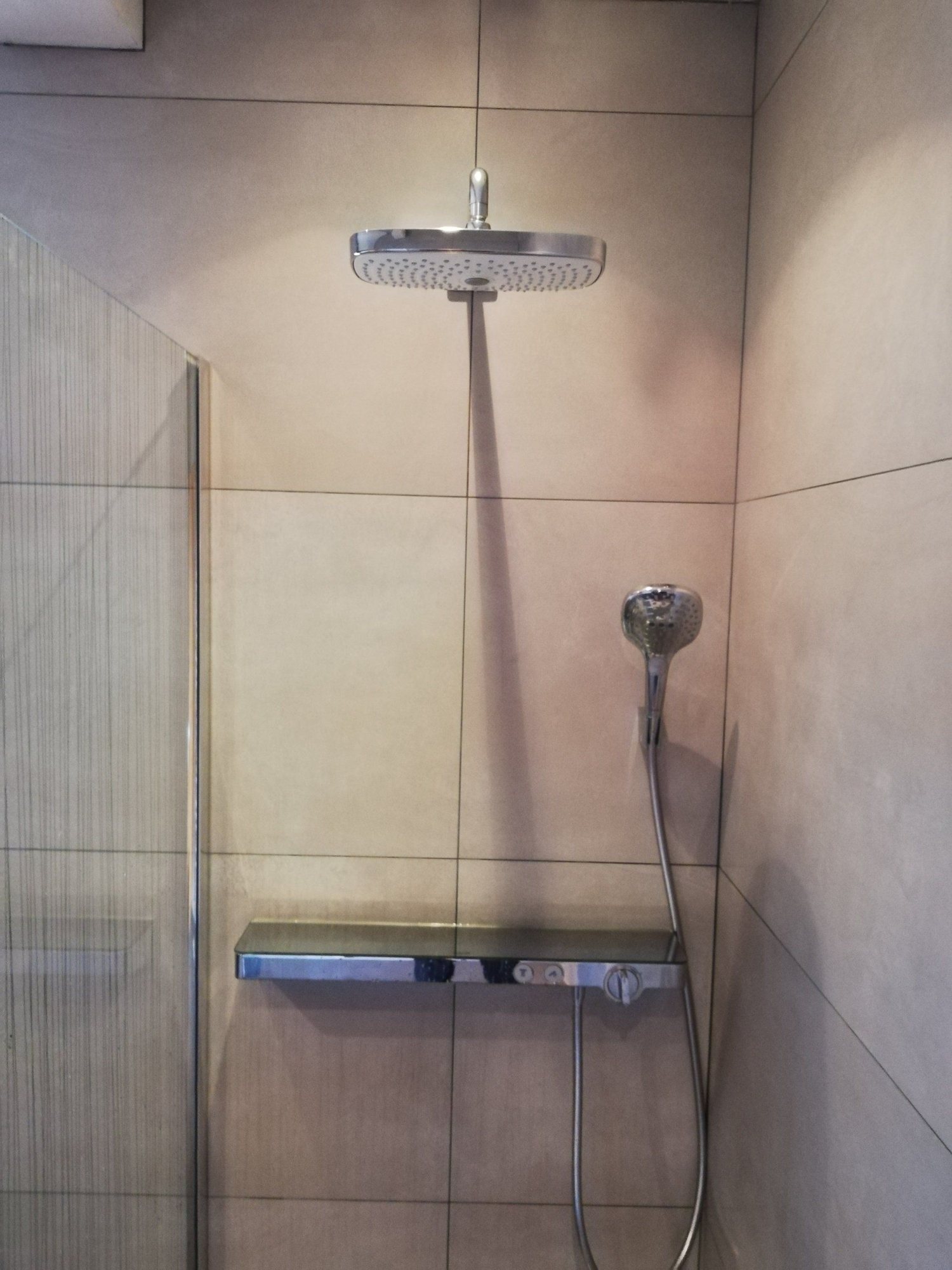 Une douche carrelée en beige équipée d'une grande pomme de douche rectangulaire, d'une douchette à main et d'une tablette thermostatique, avec une paroi vitrée à gauche.