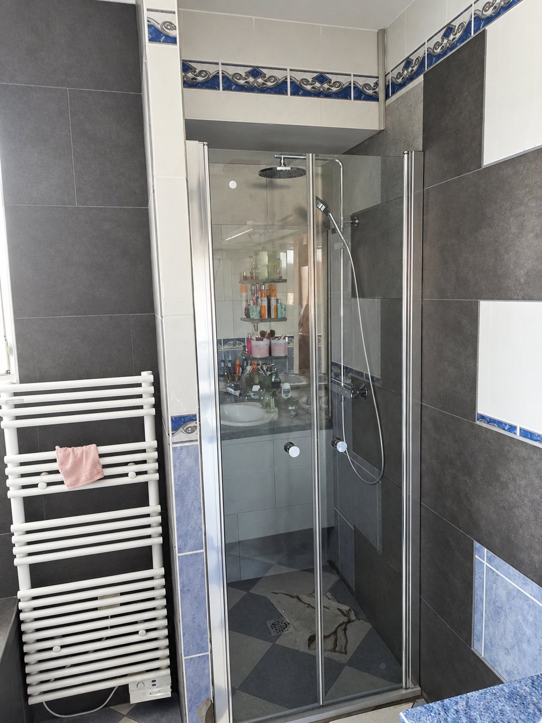 Salle de bain rénovée avec une douche fermée par une porte en verre pliante, des carreaux gris et bleus et un radiateur sèche-serviettes blanc.