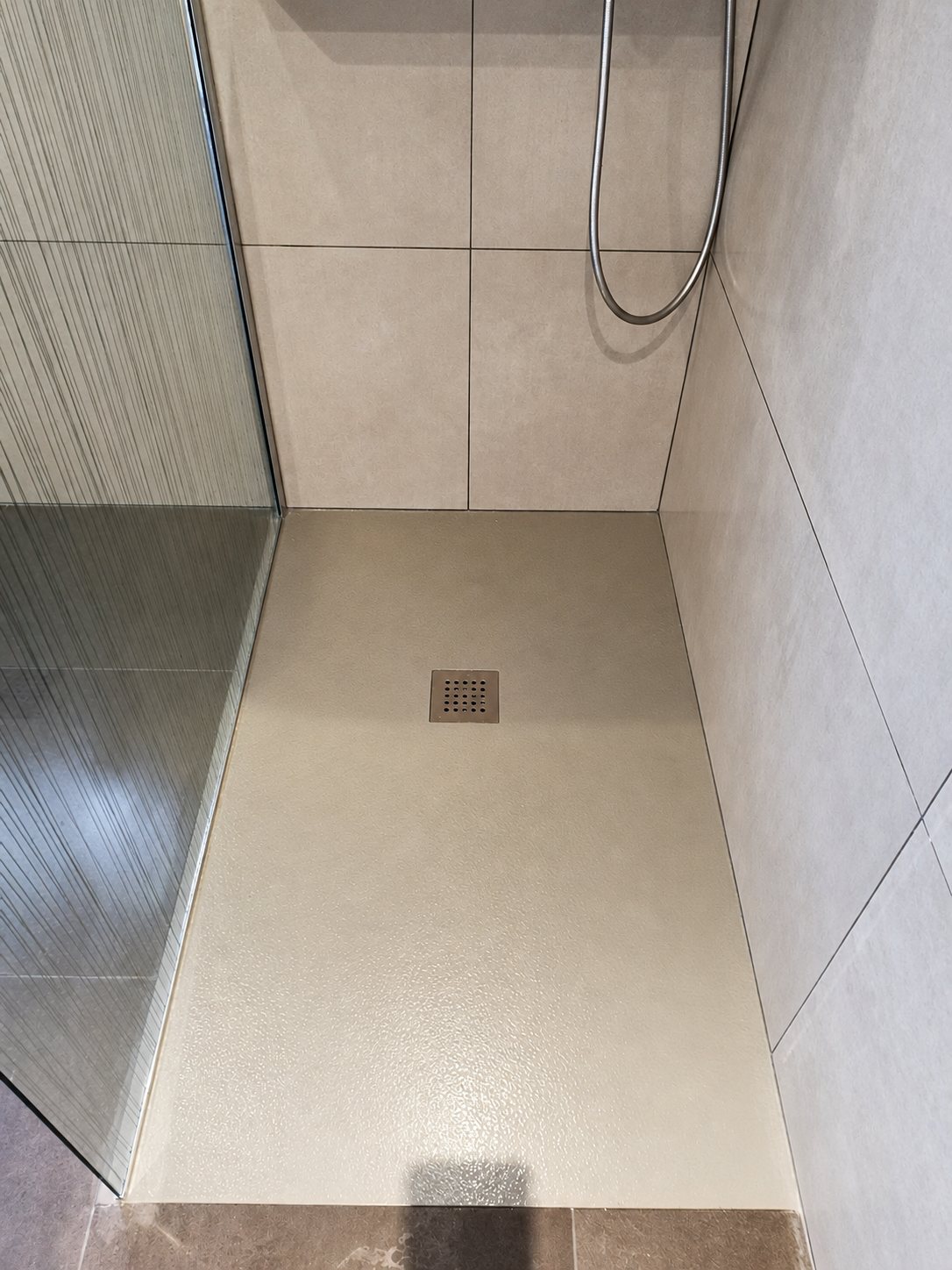Une douche à l'italienne fraîchement rénovée avec un receveur extra-plat beige, une bonde carrée en inox, une paroi vitrée et un carrelage mural gris beige.