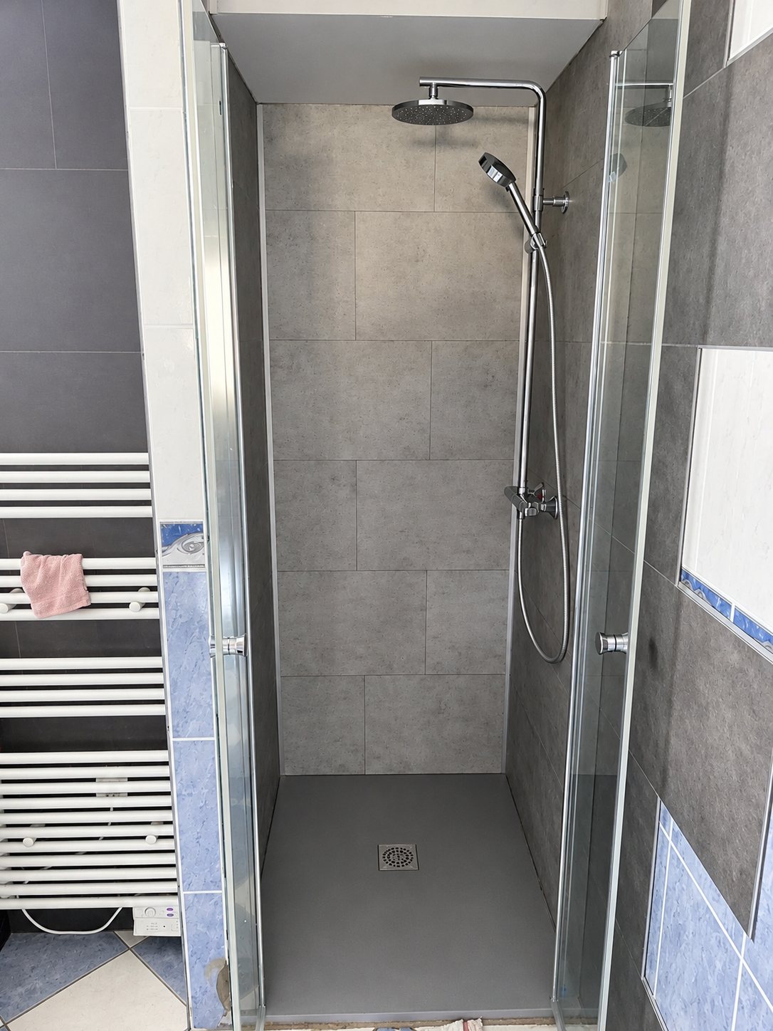 Une douche à l'italienne aux murs et au receveur gris, équipée d'une pomme de douche ronde et d'une douchette, fermée par une porte vitrée, à côté d'un sèche-serviettes blanc.