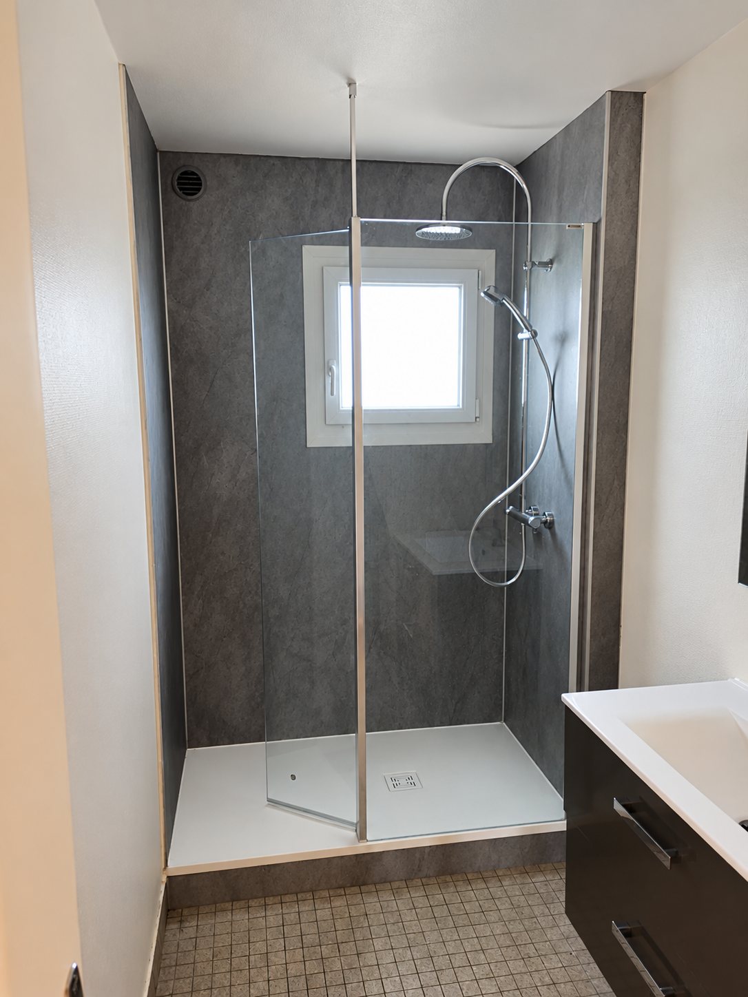 Salle de bain rénovée avec une douche à l'italienne aux parois grises effet pierre, une paroi vitrée et un meuble vasque au premier plan.