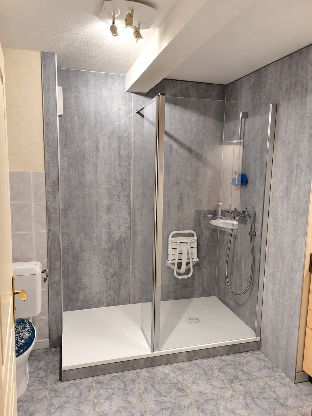 Salle de bain rénovée avec une grande douche à receveur blanc, paroi en verre, panneaux muraux gris effet béton et siège de douche rabattable blanc.