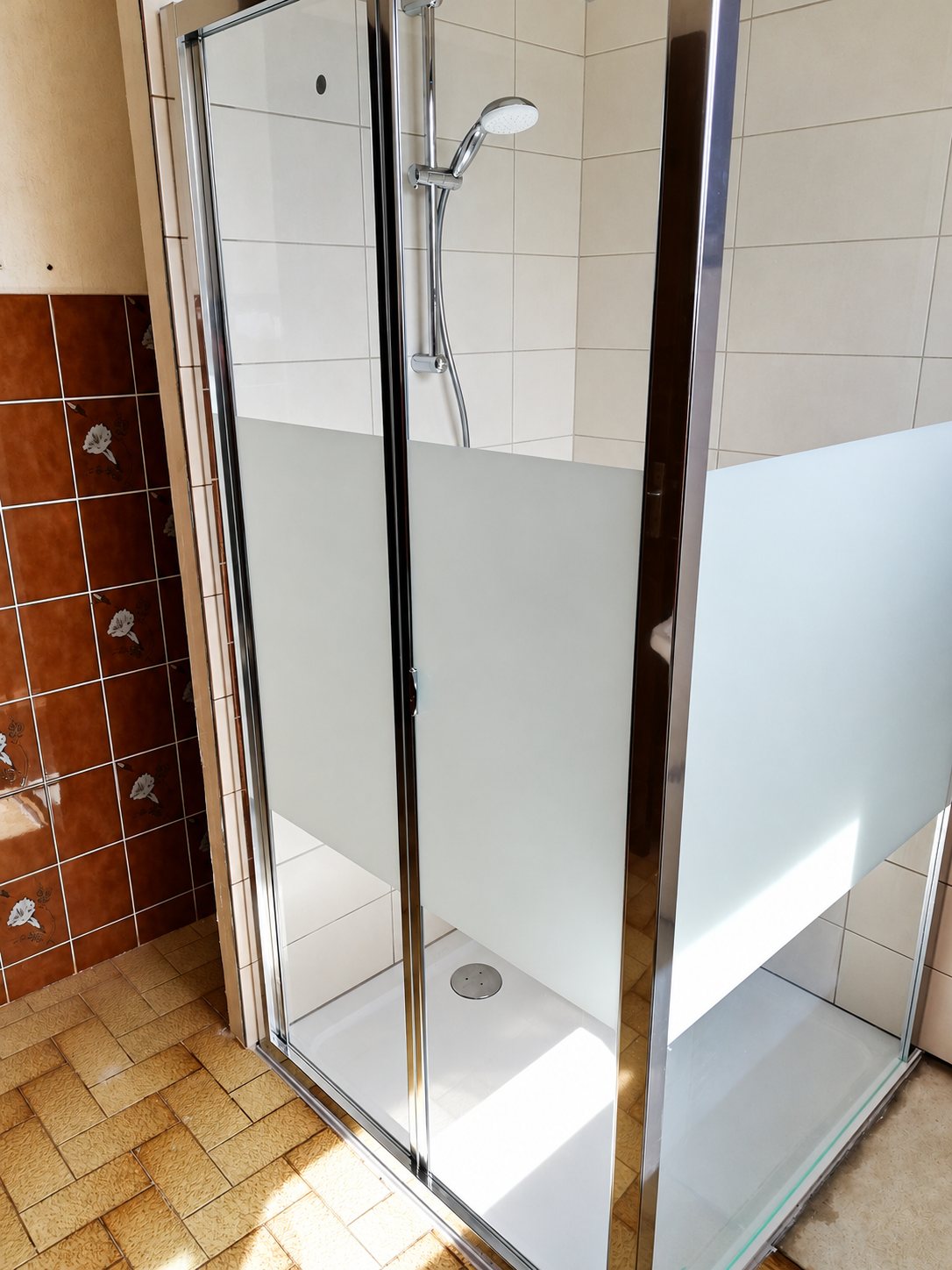 Une cabine de douche récente avec receveur blanc et parois de verre partiellement dépoli, installée dans une salle de bains aux anciens carrelages marron.
