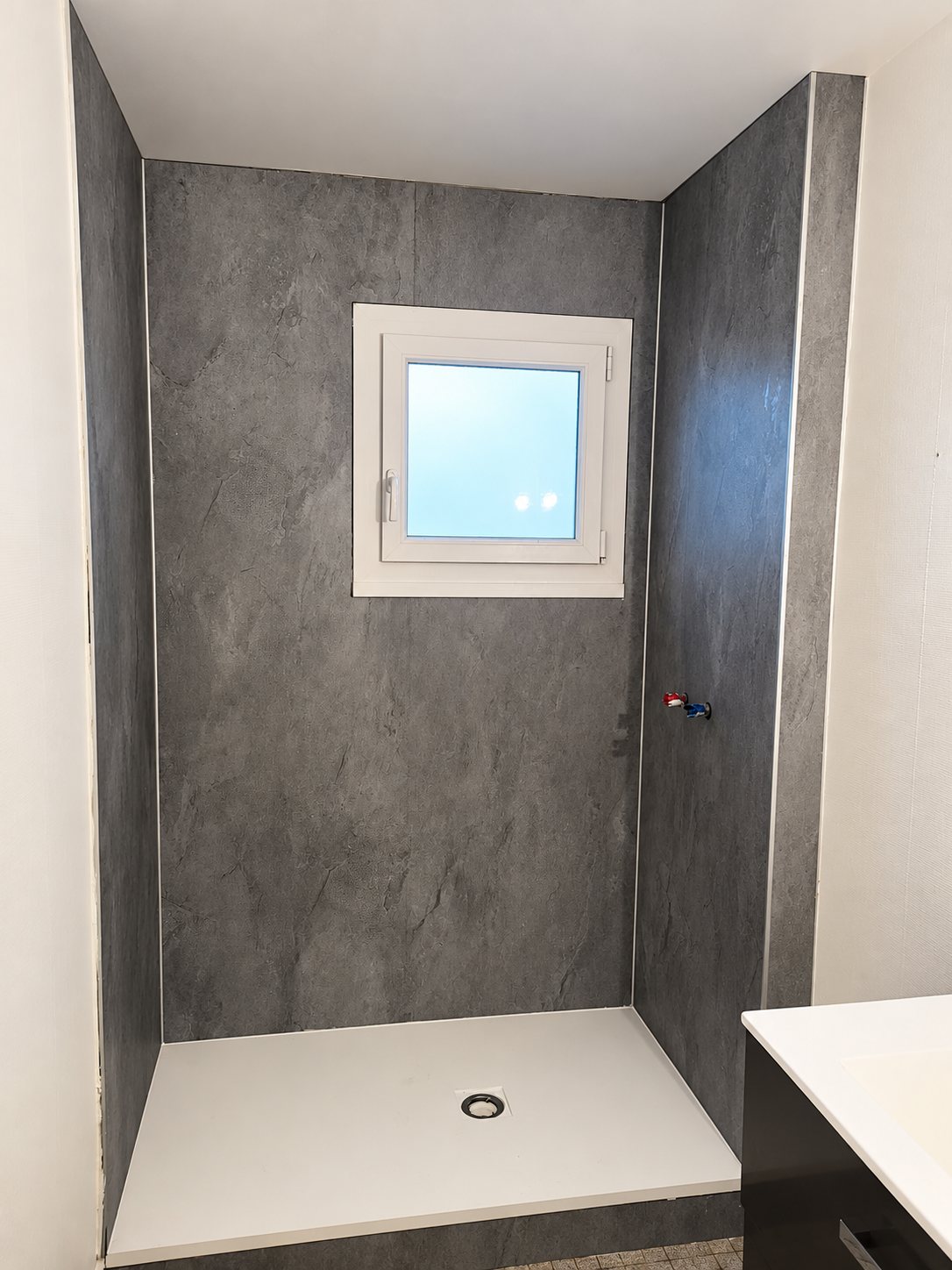 Une douche à l'italienne rénovée avec des panneaux muraux gris effet pierre, un receveur blanc extra-plat et une petite fenêtre blanche.