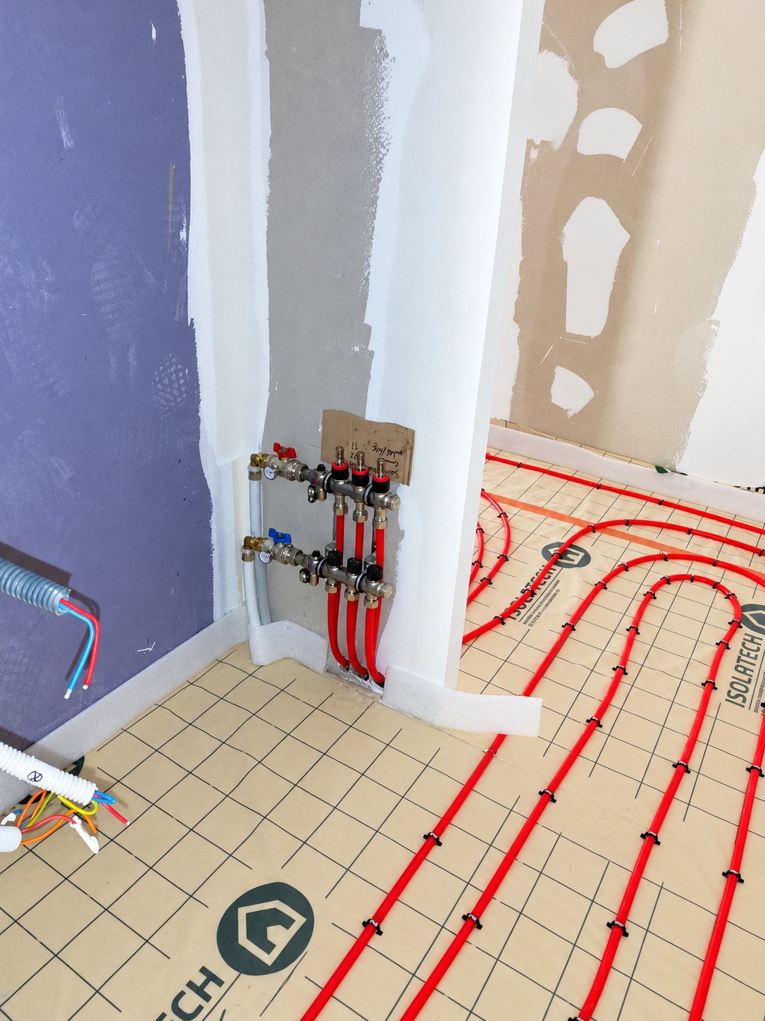 Chantier de rénovation montrant un collecteur de chauffage au sol relié à des tubes rouges posés sur un isolant quadrillé de marque Isolatech.
