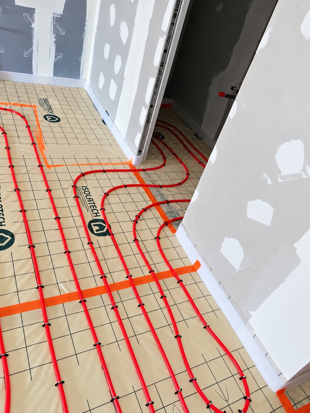 Des tubes rouges de plancher chauffant sont fixés sur une membrane Isolatech quadrillée, dans une pièce aux cloisons en plaques de plâtre.