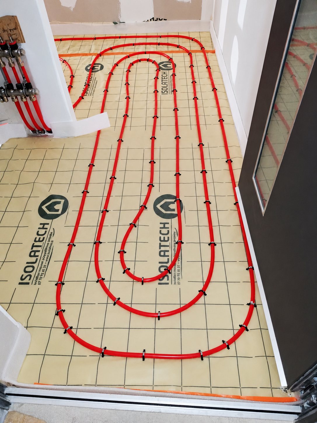 Tuyaux rouges de plancher chauffant posés en boucles sur un film isolant quadrillé Isolatech, relié à un collecteur dans une pièce en travaux.
