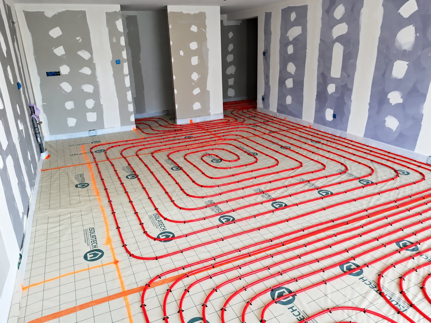 Réseau de tuyaux rouges de plancher chauffant posés en spirale sur un film isolant quadrillé dans une pièce aux murs en plaques de plâtre en cours de finition.