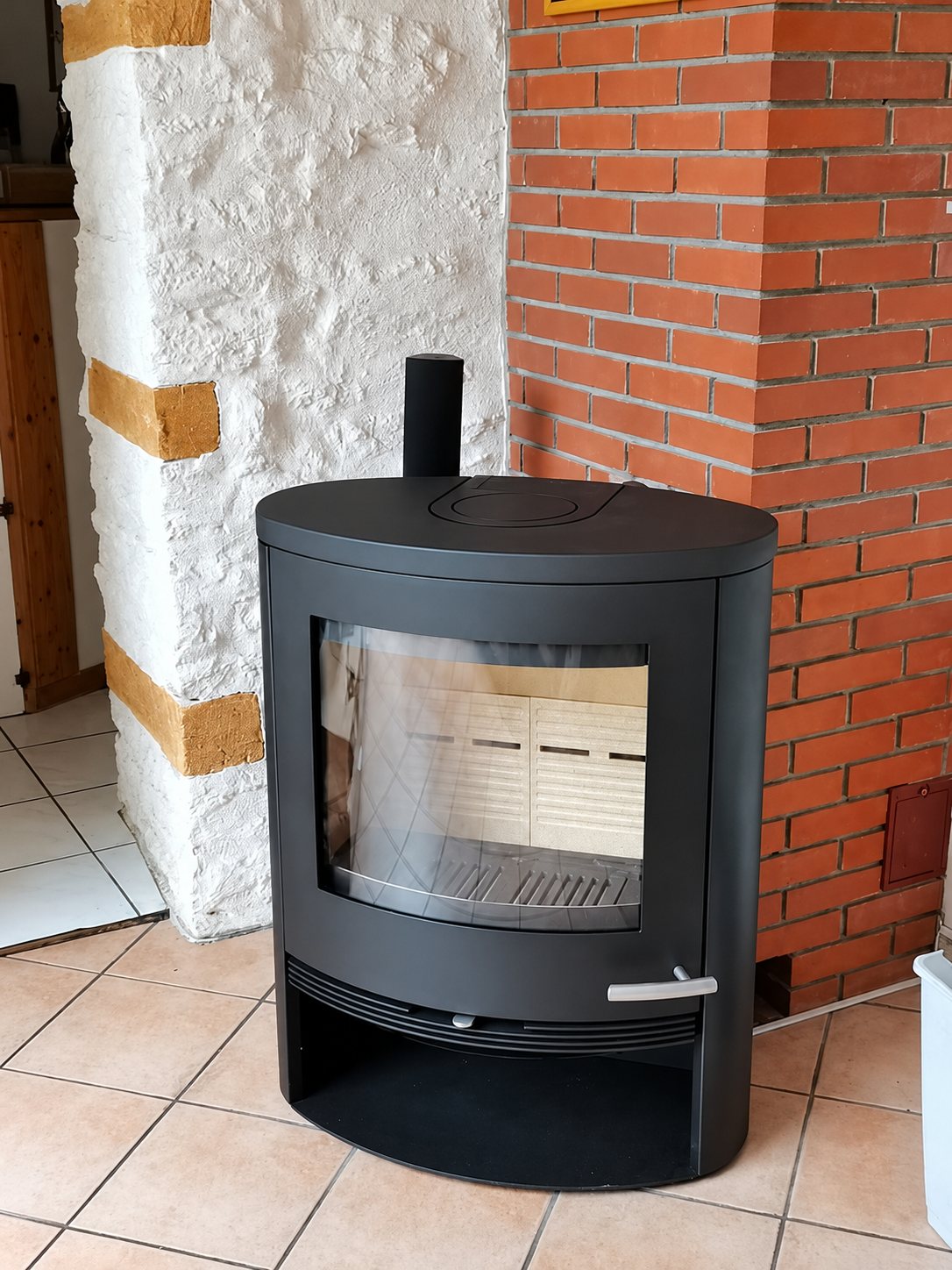 Un poêle à bois noir de forme ovale avec une grande vitre est installé sur un carrelage, entre un mur en pierre blanche et un mur en briques rouges.
