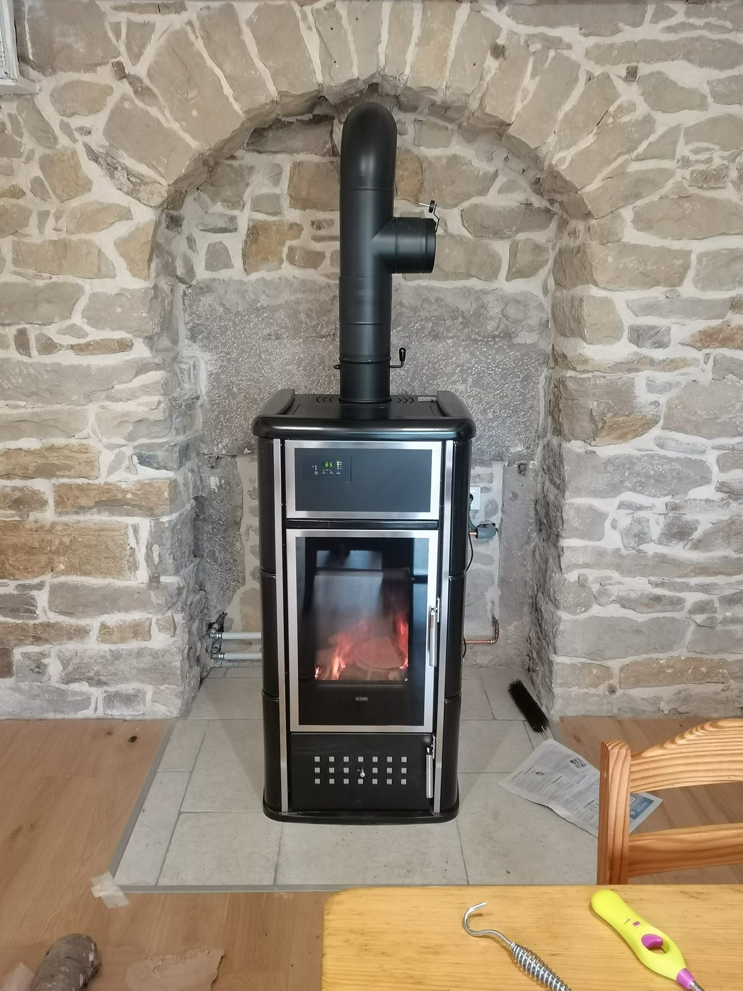 Un poêle à bois noir avec une vitre et un feu allumé est installé dans une niche en pierres apparentes, sur un sol en dalles claires.