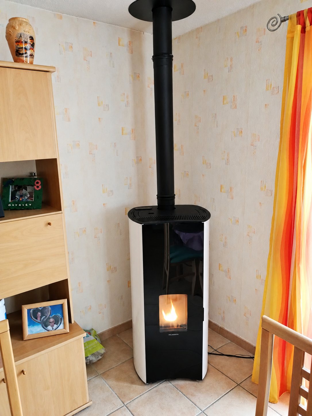 Un poêle à granulés blanc et noir avec un conduit noir est installé dans le coin d'un salon, à côté d'un meuble en bois clair et d'un rideau orange.