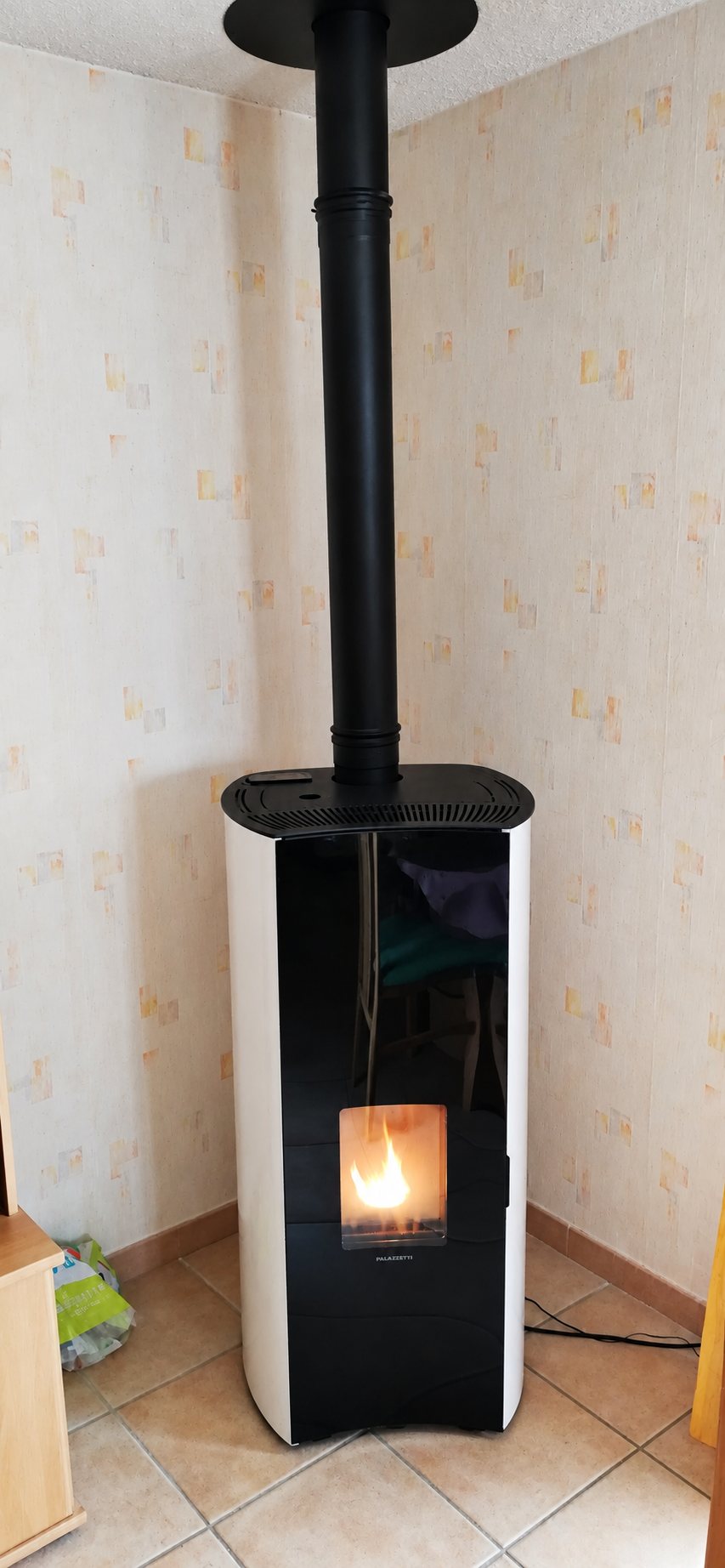 Un poêle à granulés blanc et noir, flamme allumée, est installé dans l'angle d'une pièce avec un conduit noir qui monte au plafond.