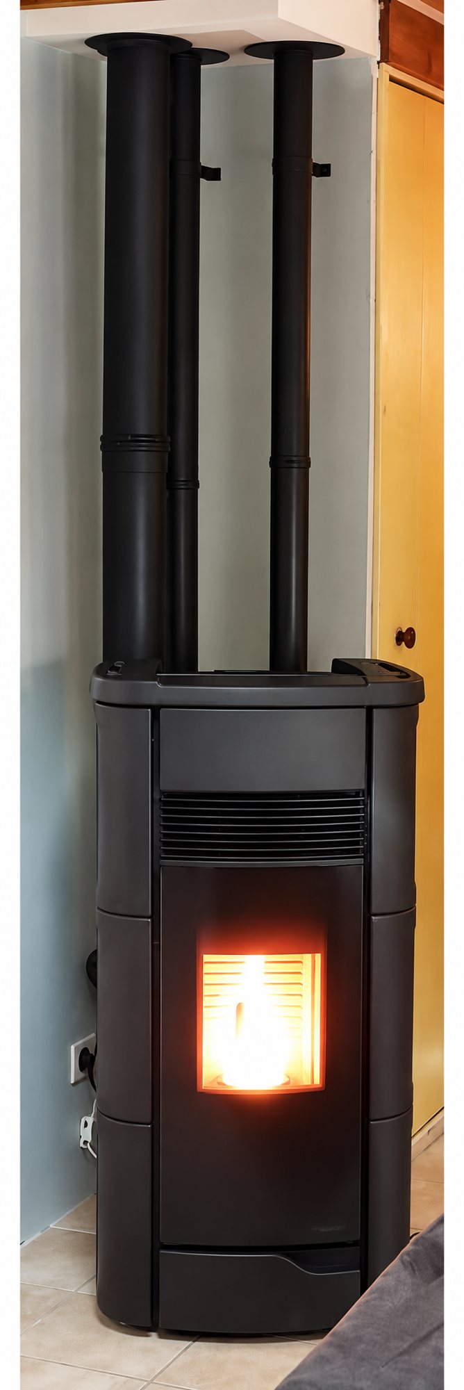 Un poêle à granulés noir avec flamme allumée, surmonté de trois conduits de fumée noirs, est installé dans un coin de pièce près d'une porte en bois.