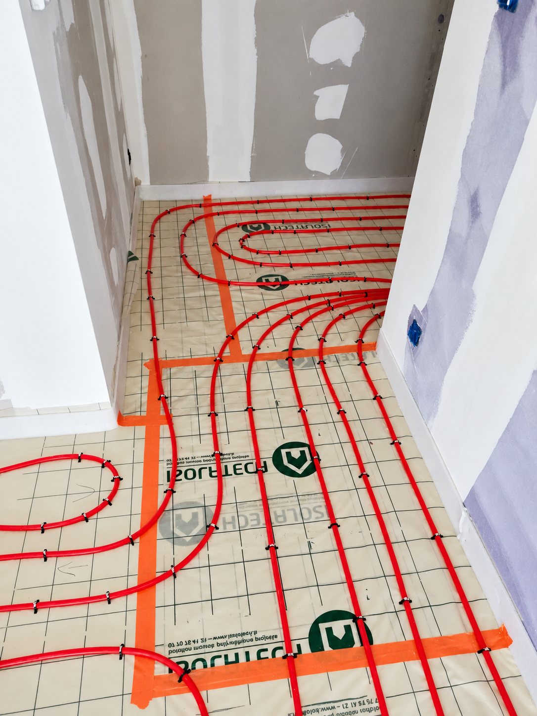 Tuyaux rouges de plancher chauffant fixés sur un film isolant Isoltech quadrillé, dans une pièce en cours de rénovation aux murs en plaques de plâtre.
