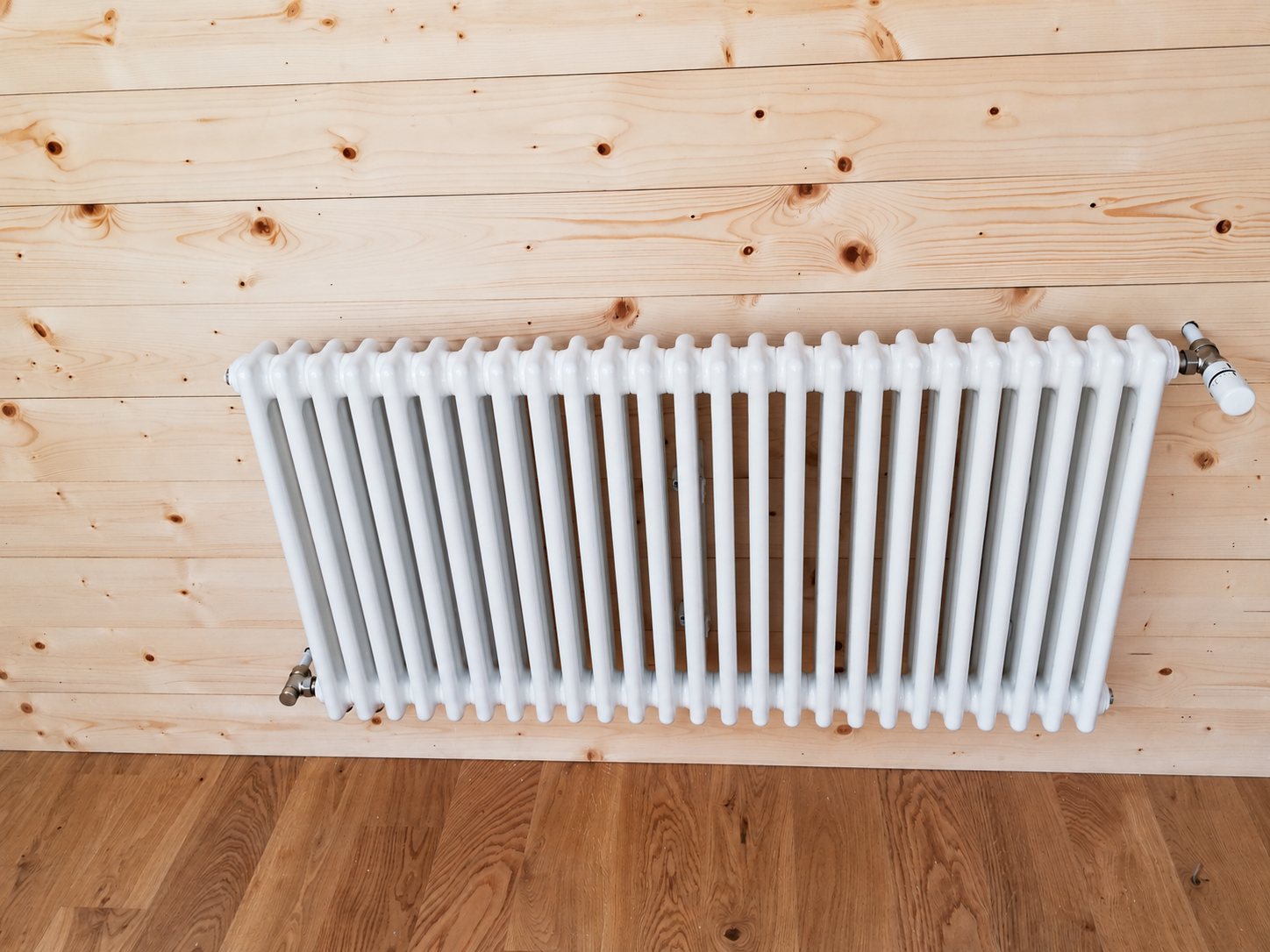 Un radiateur blanc à colonnes avec robinet thermostatique est installé sur un mur en lambris de bois clair, au-dessus d'un parquet.
