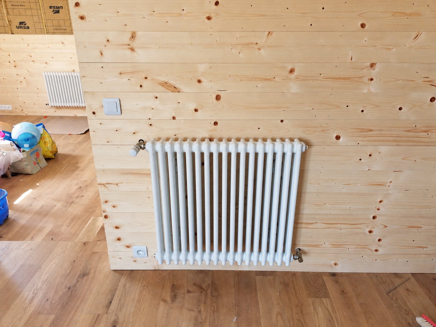 Un radiateur blanc à colonnes est fixé sur un mur en lambris de bois clair, dans une pièce en rénovation avec parquet.