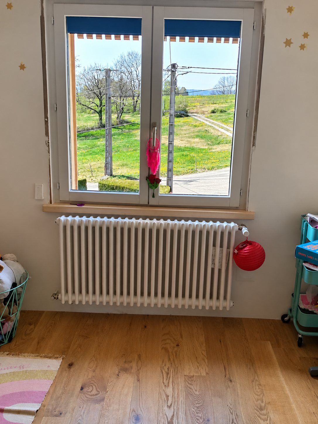 Un radiateur blanc à éléments est installé sous une fenêtre donnant sur un champ, dans une chambre avec parquet en bois.