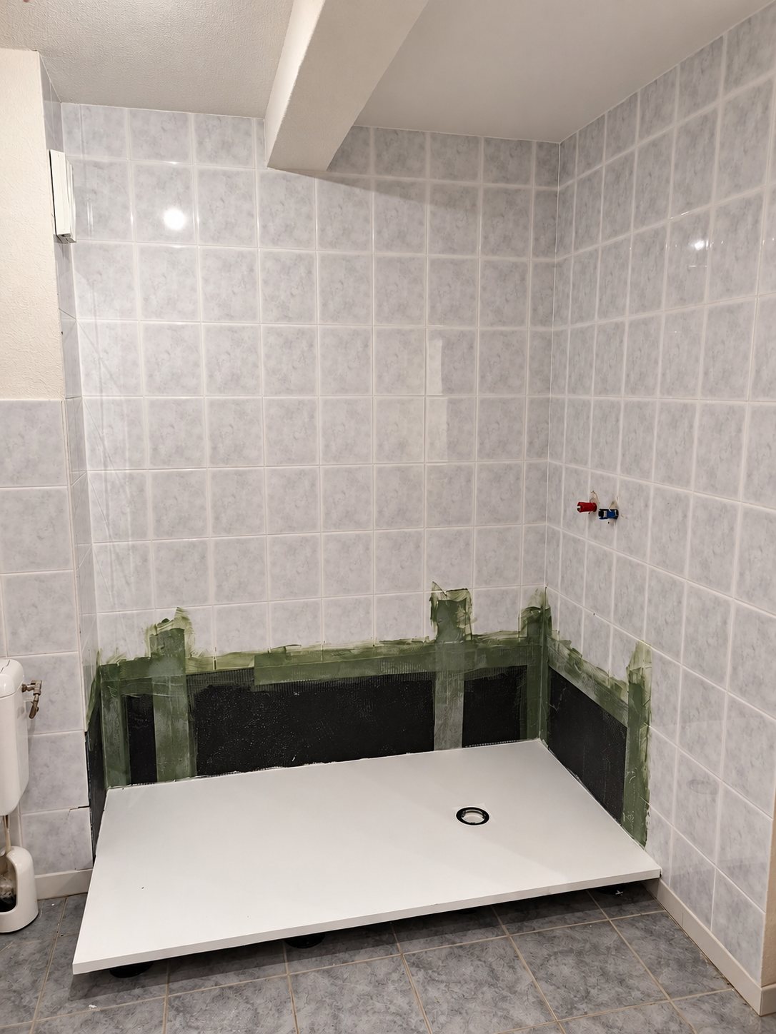 Salle de bain en travaux avec un receveur de douche blanc posé au sol et un mur carrelé dont le bas est recouvert d'une étanchéité verte.