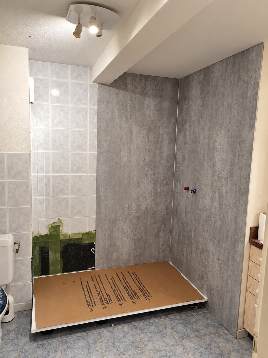 Salle de bain en cours de rénovation avec un receveur de douche protégé par du carton et des panneaux muraux gris aspect béton.