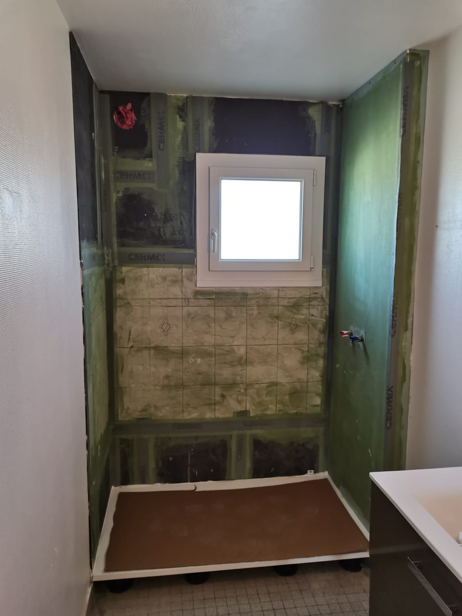 Salle de bain en cours de rénovation avec un receveur de douche installé, des panneaux de construction verts sur les murs et une petite fenêtre blanche.