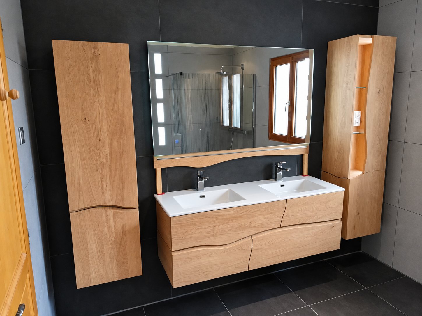 Salle de bain aux murs sombres équipée d'un meuble suspendu en chêne à double vasque blanche, d'un grand miroir lumineux et de deux colonnes de rangement en bois.