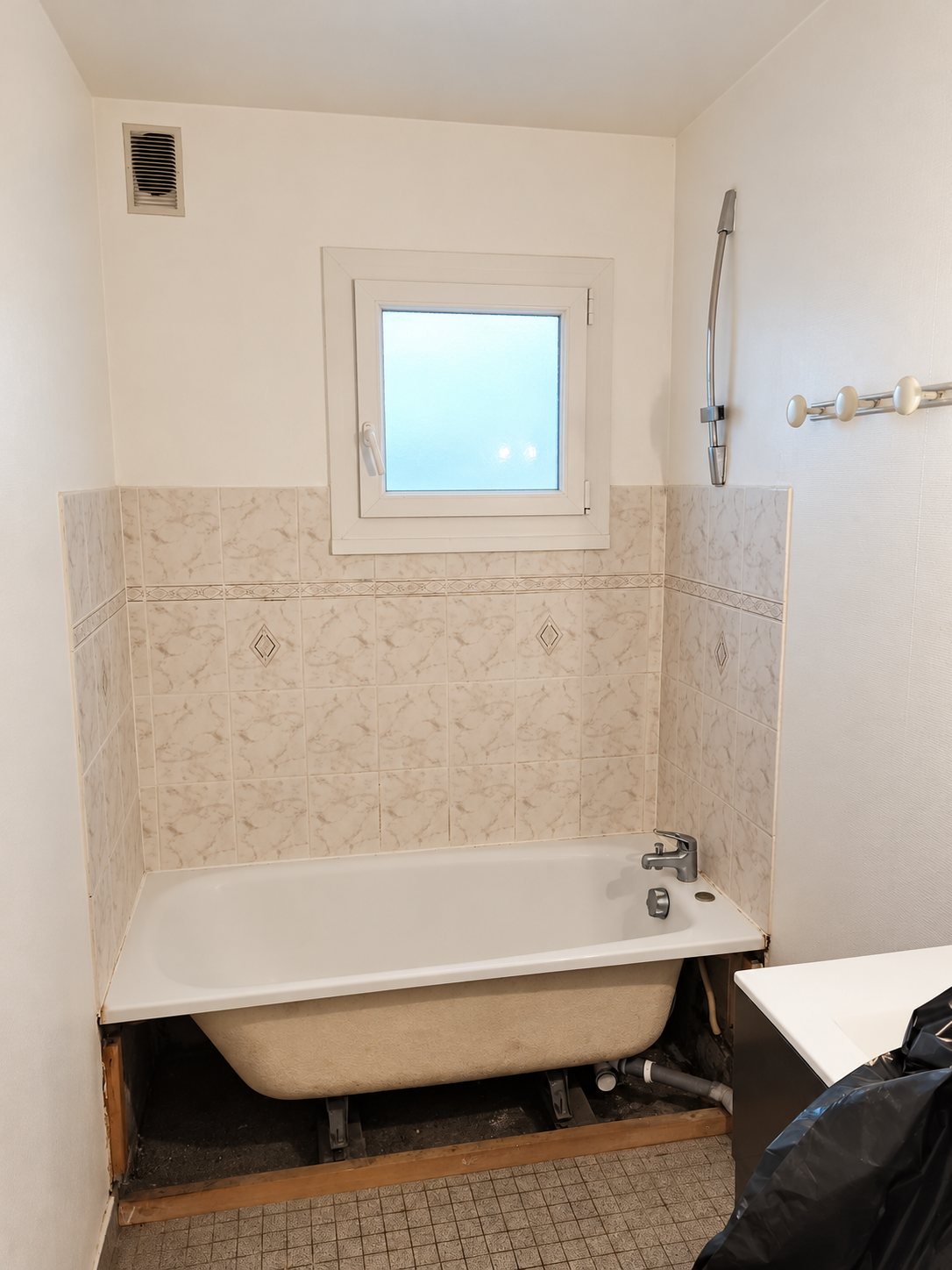 Salle de bain en travaux avec une baignoire blanche dont le tablier est retiré, un carrelage beige marbré et une petite fenêtre.