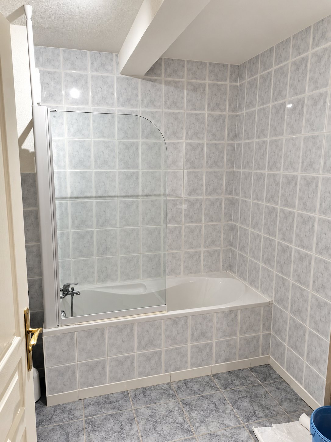 Salle de bain carrelée de gris clair avec une baignoire équipée d'un pare-baignoire en verre et une porte blanche à poignée dorée.