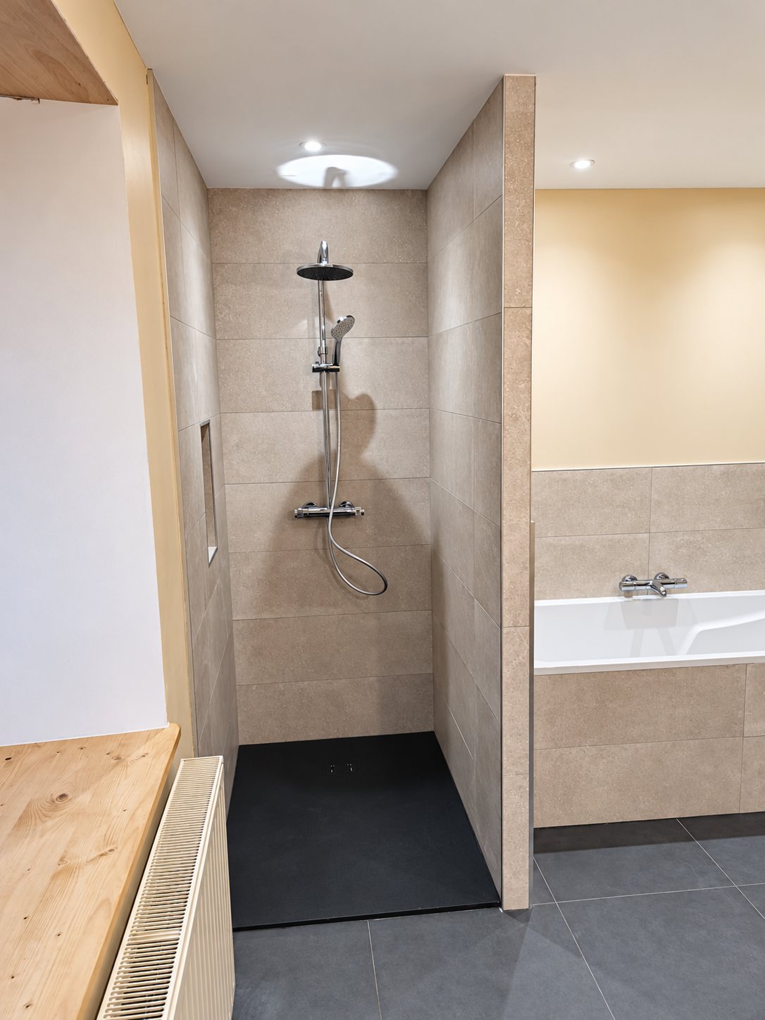 Salle de bain rénovée avec une douche à l'italienne carrelée de beige, un receveur anthracite, une baignoire blanche et un radiateur au premier plan.