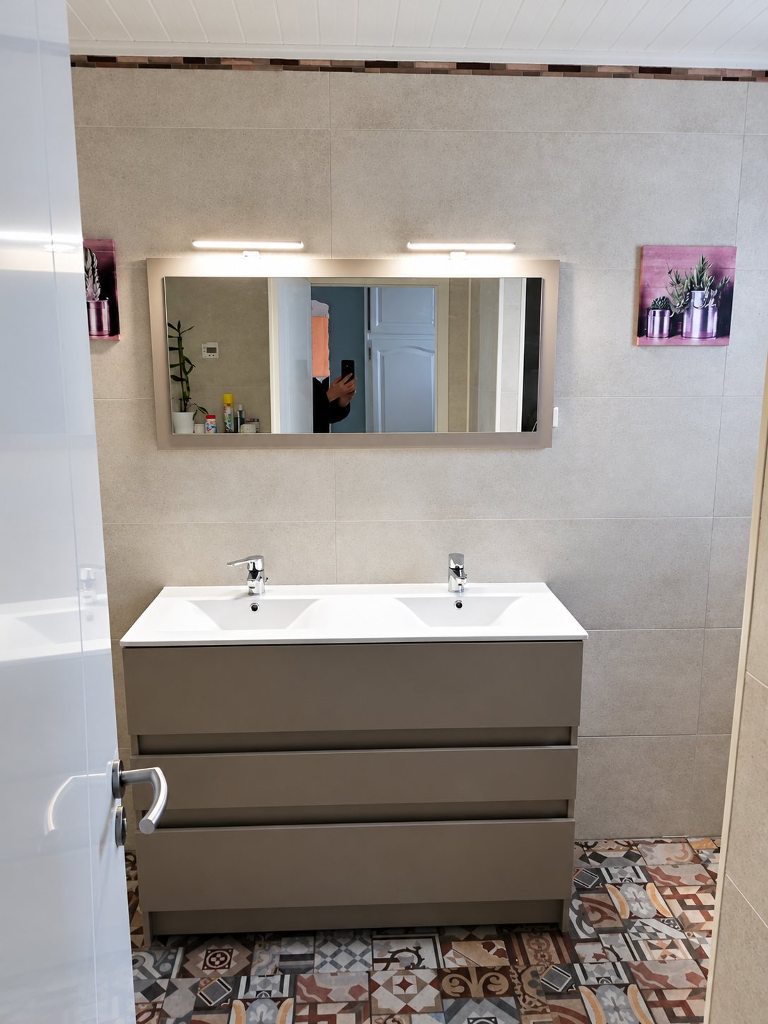 Salle de bain rénovée avec un meuble gris à trois tiroirs et double vasque blanche, un grand miroir éclairé et un sol en carreaux de ciment colorés.