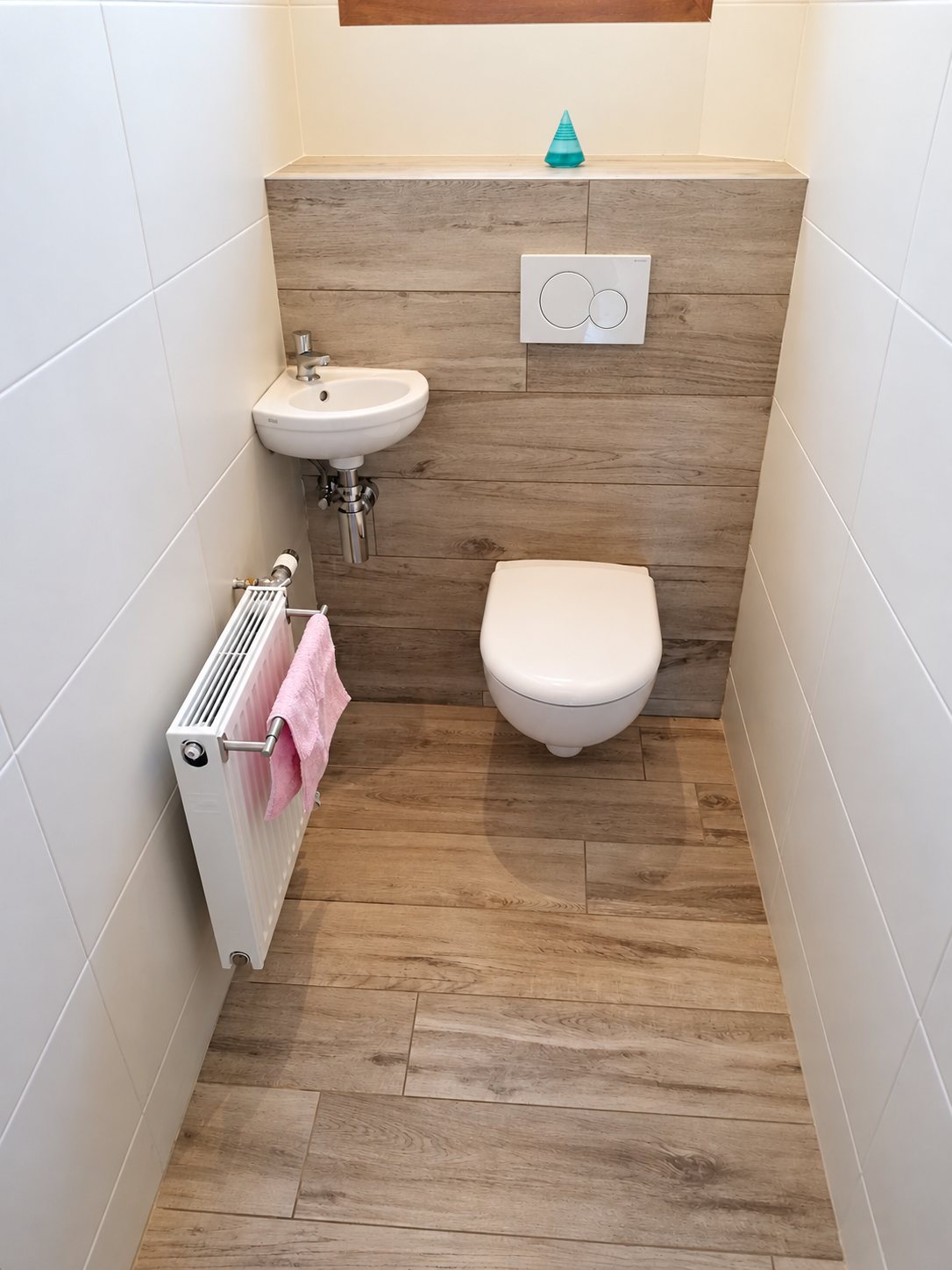 Petites toilettes rénovées avec WC suspendu, lave-mains d'angle, radiateur et carrelage au sol et au mur imitation bois.