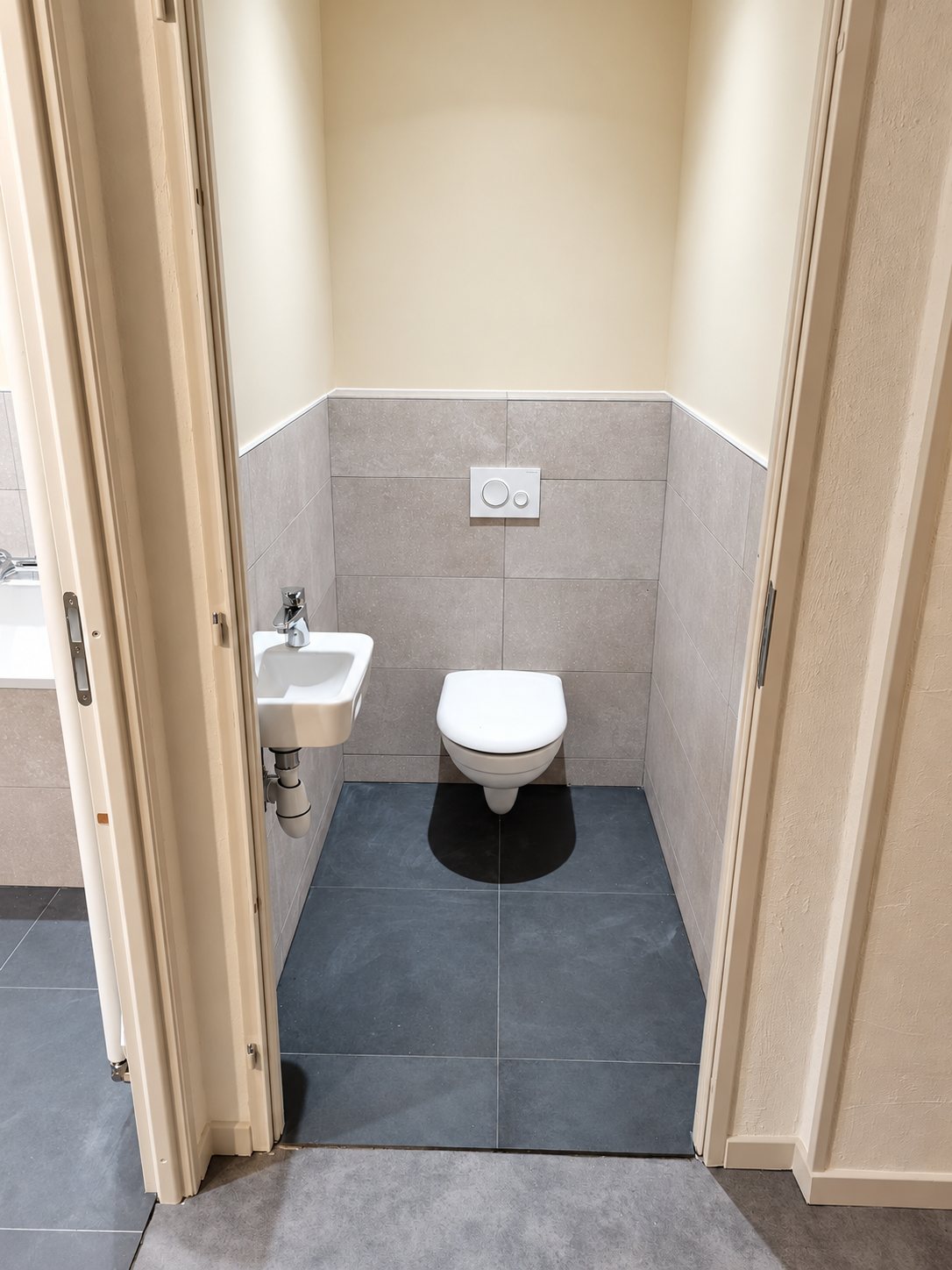 Petit WC rénové avec cuvette suspendue blanche, lave-mains d'angle, carrelage gris au sol et sur les murs, vu depuis l'entrée.